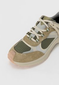 Vihreät ja beige kengät, joissa on sekoitettuja materiaaleja, kuten mokkaa ja verkkoa, paksut nauhat ja teksturoitu, pehmustettu pohja. Huomattava ruutukuvio sivussa.