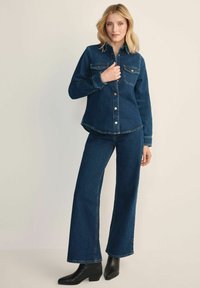 Kobieta stoi w ciemnoniebieskiej koszuli dżinsowej i szerokich dżinsach, w czarnych botkach za kostkę, pozując na neutralnym tle.