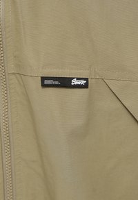 Tissu beige avec une étiquette noire portant l'inscription « STAY UNITED DESIGN IN EUROPE » et des coordonnées cousues près d'une fermeture éclair verticale.