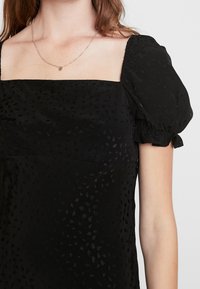 Musta puff-sleeve-mekko neliön muotoisella pääntiellä. Teksturoitu kangas sisältää hienovaraisia pilkkuja. Lyhyet hihat on kerätty hihansuissa.