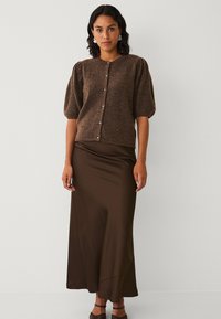 Bruine gebreide cardigan met korte pofmouwen, parelknoopjes, gecombineerd met een satijnen bruine maxi rok. Soepele textuur en elegante pasvorm.