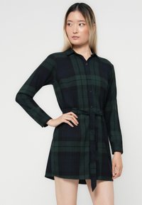 Robe chemise en flanelle à carreaux verts foncés et bleus marins. Dotée d'un col, de manches longues, d'une fermeture à boutons à l'avant et d'une ceinture assortie à la taille.