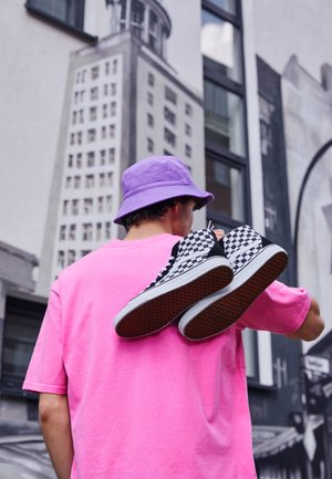 Personne portant un chapeau seau violet et une chemise rose, transportant des baskets à carreaux noirs et blancs sur l'épaule, avec des bâtiments urbains en arrière-plan.