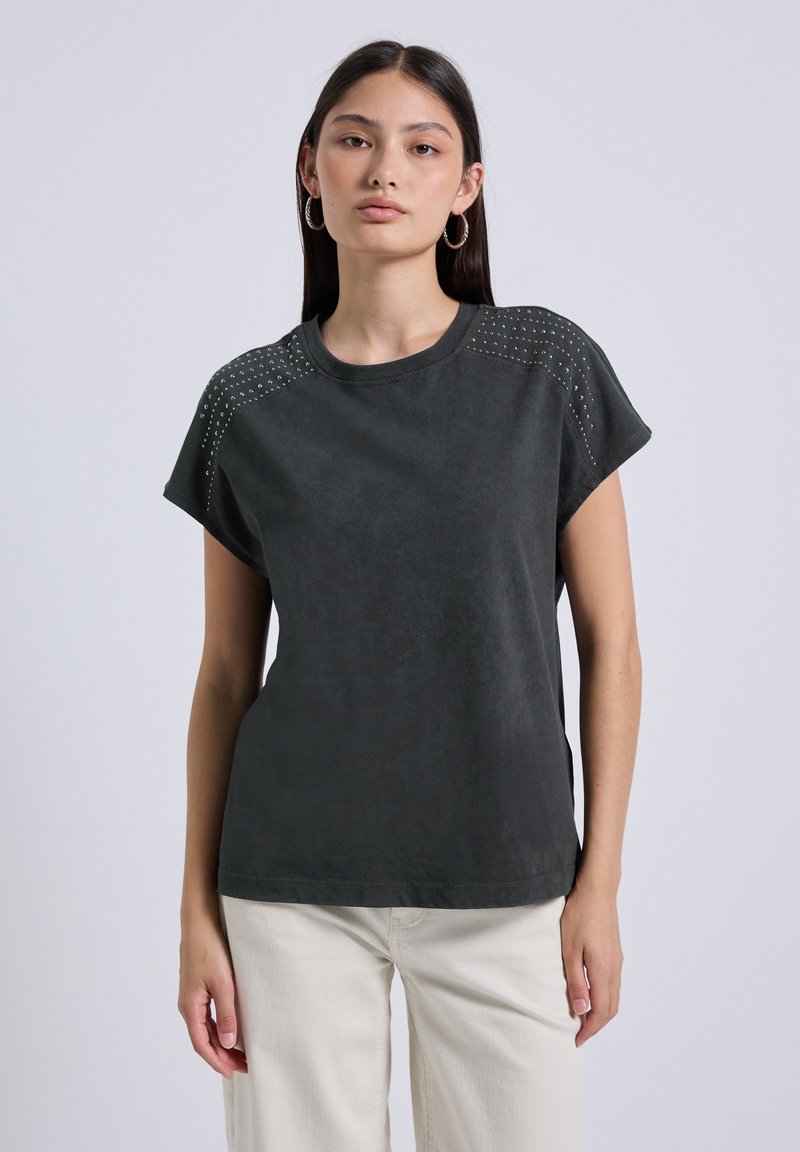Camiseta de manga corta gris oscuro con tejido texturizado, acentos de tachuelas en los hombros y escote redondo. Se combina con pantalones beige claros.