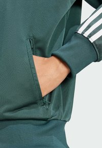 Green athletic jacket with ribbed cuffs, featuring a side pocket and white striped accents on the sleeve. Smooth texture, fitted design.
