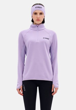 Vrouw draagt een lichtpaarse lange mouwen top met rits en bijpassende hoofdband, met zwarte leggings, staand tegen een effen achtergrond.