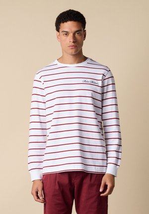 Junger Mann mit weißem Langarmshirt mit roten Querstreifen und dunkelroter Hose steht vor einem schlichten beigen Hintergrund.