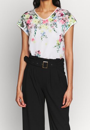 Femme portant un top blanc sans manches à motif floral avec des accents roses, jaunes et verts, associé à un pantalon noir taille haute avec ceinture.