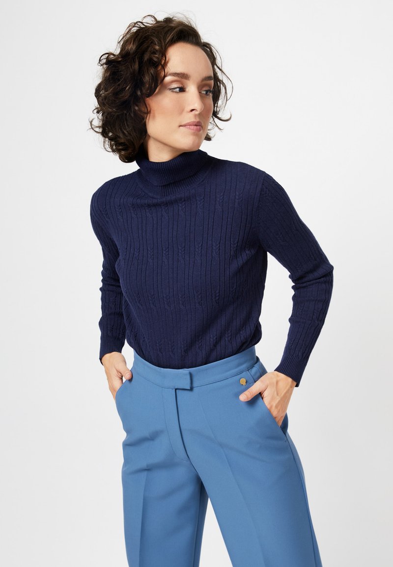 Pull en laine côtelée bleu marine avec col roulé, associé à un pantalon ajusté bleu clair comportant des poches latérales et une finition en bouton doré.