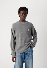 Junger Mann mit kurzen Dreadlocks, der einen grauen Strickpullover mit einem kleinen Logo und weit geschnittenen blauen Jeans trägt, steht vor einem einfarbigen weißen Hintergrund.