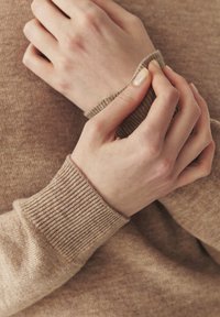 Pull beige en maille avec poignets côtelés, mains ajustant la manche. Le matériau semble doux et chaud, avec une texture subtile.