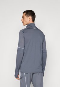 Grå atletisk jakke med høy krage, lange ermer og hvite kontrastsømmer, laget av glatt, lett stoff. Under Armour-logo på baksiden.