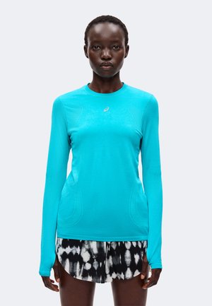 Femme portant un maillot de sport bleu vif à manches longues et un short à motifs noir et blanc, debout devant un fond uni.
