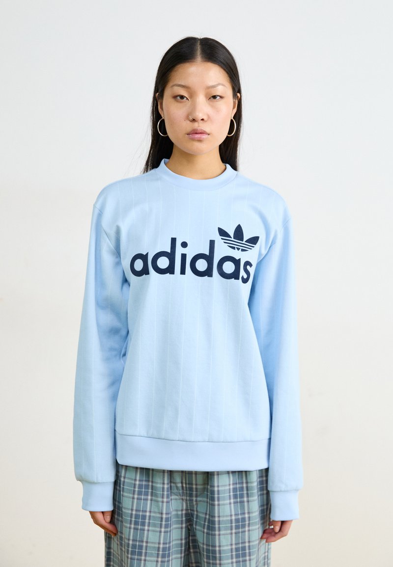 Gaiši zila Adidas sporta jaka ar garām piedurknēm, ribota struktūra un liels melns logo. Kombinēta ar rūtainām biksēm zilās un zaļās krāsās.
