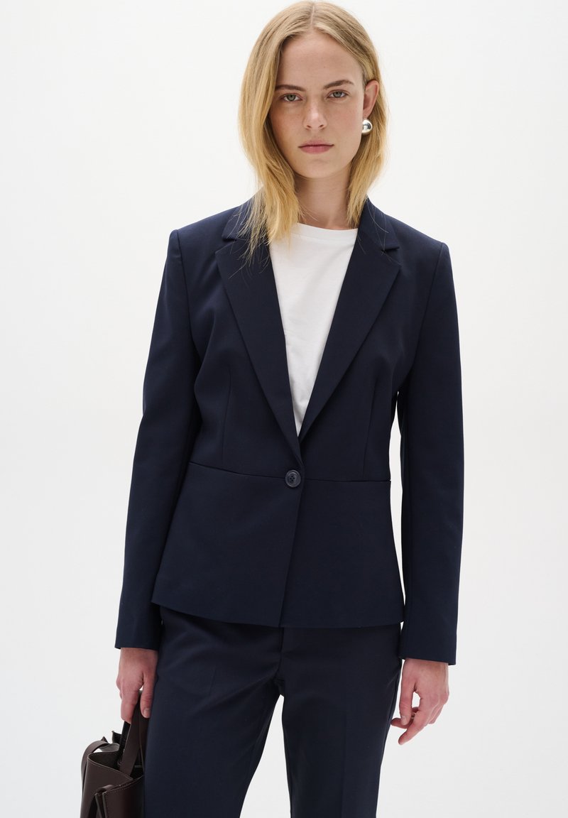 Námořnický blazer s jedním knoflíkem, přizpůsobený design, klopové límce a hladká textura. Kombinován s bílou košilí a tmavou kabelkou.