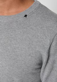 Gros plan d'une personne portant un pull tricoté gris clair avec un petit logo noir brodé près du col.