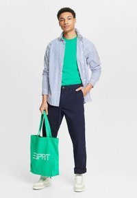 Jauns vīrietis valkā svītrainu krekls, zaļu T-kreklu, tumši zilos bikses, baltas sporta kurpes, tur zaļu somu ar 'ESPRIT' logo.