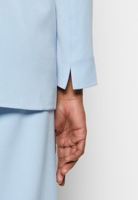 Blusa entallada de un azul claro con un primer plano de una manga que presenta un detalle de abertura, mostrando una textura suave y líneas limpias, con una mano posicionada cerca.