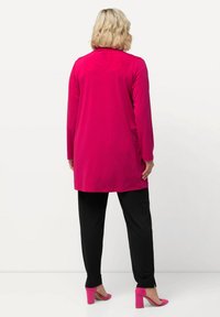 Fuchsia-langärmlige Tunika, kombiniert mit schwarzen, schmalen Hosen. Rosa High Heels setzen einen Farbakzent zum Outfit. Weicher Textur sichtbar.