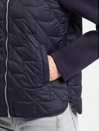 Marineblauer, gesteppten Pufferweste mit geometrischer Naht, silbernem Reißverschluss und seitlicher Tasche. Erhöhte Kragen und kontrastierendes Ärmelmaterial.