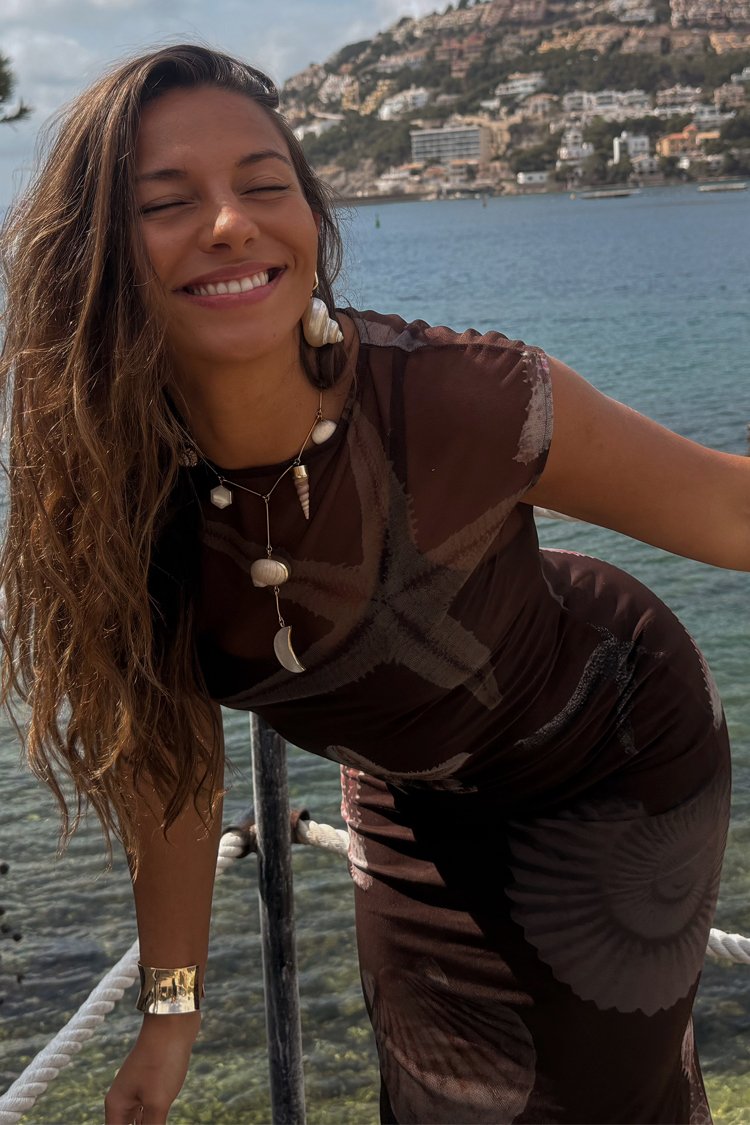 Brązowa sukienka w wzory z motywem rozgwiazdy; modelka nosi biżuterię w formie muszli i półksiężyca; plażowa sceneria z czystą wodą w tle.