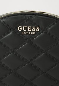 Pochette in pelle ecopelle nera trapuntata con logo dorato "GUESS EST 1981". Caratterizzata da una parte superiore arrotondata e cuciture a contrasto per un tocco di dettaglio.