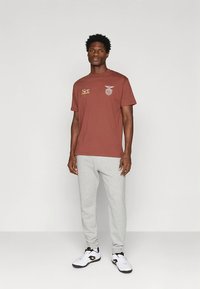 Bruin grafisch T-shirt met korte mouwen, met een embleem en tekst. Gecombineerd met lichtgrijze joggingbroek en witte sportschoenen.