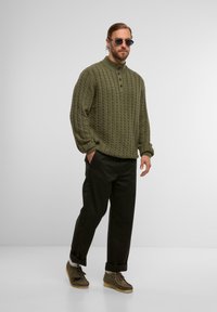 Olijfgroene kabelgebreide trui met een hoge kraag en drie knopen, gecombineerd met een donkere broek en bruin veterschoenen. Accessoires zijn onder andere een zonnebril.