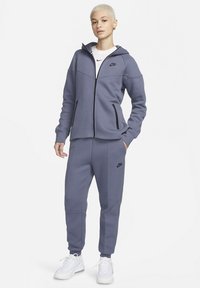 Grijze Nike trainingspak met een rits hoodie en voork pockets, gecombineerd met bijpassende broek. Het materiaal lijkt zacht en rekbaar met een slim fit ontwerp.