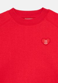 Rdeča majica s dolgimi rokavi in krožnim ovratnikom ter rebrastimi manšetami, ki ima majhen vezen srčkast značek z napisom "KLUB V.I.P.".