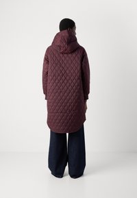 Lange gewatteerde jas in diep bordeauxrood met een capuchon. Heeft een diamondpatroon en elastische manchetten. Draag het met losse, donkere denimbroeken.
