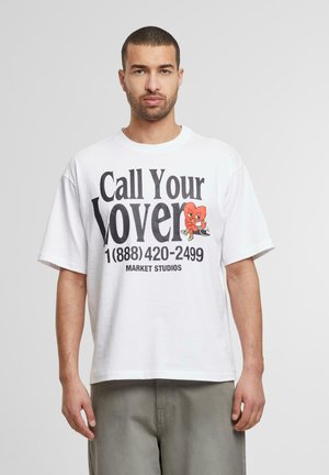 Hvid bomuld t-shirt med oversized pasform, der har fed sort tekst "Ring til din elskede" og et tegneserie-hjerte design. Korte ærmer, rund hals.