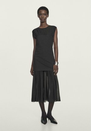 Schwarzes Kleid mit enganliegendem Oberteil und ausgestelltem, halbtransparentem Rock. Kurze Flügelärmel und strukturierte Details an der Taille. Trägt schwarze Schuhe.
