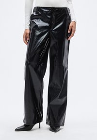 Μαύροι ψεύτικοι δερμάτινοι παντελόνες με φαρδιά γραμμή και γυαλιστερό φινίρισμα, με κλείσιμο με κουμπί μπροστά και πλαϊνές τσέπες. Συνοδεύονται από μυτερές ψηλές γόβες.