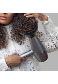 Φυσιτήρας μαλλιών Remington με κομψό γκρι σώμα και μαύρη προσαρμογή διαχύτη. Χρήστης με στυλιζαρισμένα σγουρά μαλλιά και λευκή μπλούζα με κεντητό σχέδιο.