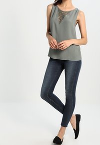 Haut sans manches vert clair avec une insertion en dentelle en forme de V ; associé à un jean skinny bleu foncé et des chaussures noires sans lacets.