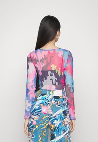 Camisola de manga longa, translúcida, com um padrão ondulado multicolorido em rosa, azul e púrpura, combinada com calças azuis estampadas que apresentam desenhos gráficos.
