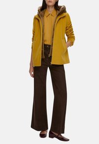 Giacca gialla con zip e cappuccio, indossata sopra una maglietta senape e pantaloni larghi marroni. L'outfit presenta linee semplici e una minimalità negli accessori.