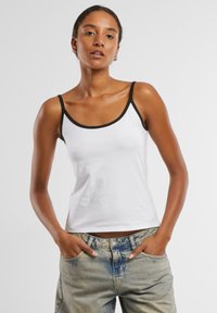 Hvit tanktop med svart kant, med tynne stropper og en tettsittende design. Båret med lyseblå denimjeans, med hendene plassert i lommene.