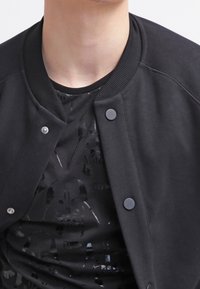 Černá bomber bunda s žebrovaným límcem a knoflíkovým zapínáním, přes černé tričko s jemným vzorovaným designem.