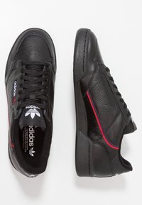 Zapatillas de cuero negras de Adidas con franjas rojas y azules, cordones negros y logo blanco en la lengüeta y el costado, mostradas desde vistas superior y lateral.