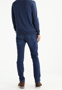 Pull bleu marine à col rond, associé à un jean bleu slim. Le jean possède des poches arrière et des détails de couture subtils. Des chaussures marron complètent la tenue.