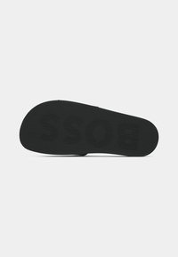 Semelle de sandale tong noire avec grip texturé et lettrage en relief du nom de la marque visible sur la surface inférieure.