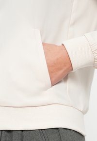 Main insérée dans la poche latérale d'un sweat-shirt crème avec poignets et ourlet côtelés, porté sur un pantalon plissé gris.