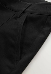 Pantalones negros con un diseño estructurado, que cuentan con un frente plano, detalles de pliegues y una textura de tela suave. Las costuras visibles acentúan los bordes.