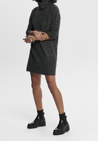 Robe en maille douce, gris foncé, avec un col roulé, des manches trois-quarts et une coupe décontractée. Associée à des bottines noires brillantes.