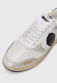 Zapatilla de cuero blanca con detalles plateados, ante texturizado, vampiro perforado, cordones blancos planos, parche redondo negro con logo y marca dorada en el lateral.