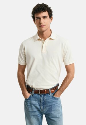 Polo shirt in crèmekleur, korte mouwen, met een kraag en een knopenlijst met twee knopen. Gekozen in combinatie met lichtblauwe jeans en een bruine riem.