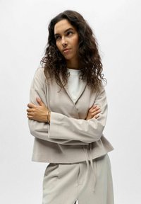 Femme aux cheveux bouclés portant un costume beige et une chemise blanche, les bras croisés, regardant légèrement sur le côté sur un fond uni.