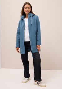 Femme portant une veste en polaire bleu clair zippée, un t-shirt blanc, un pantalon noir et des baskets blanches avec des accents noirs, se tenant devant un fond neutre.